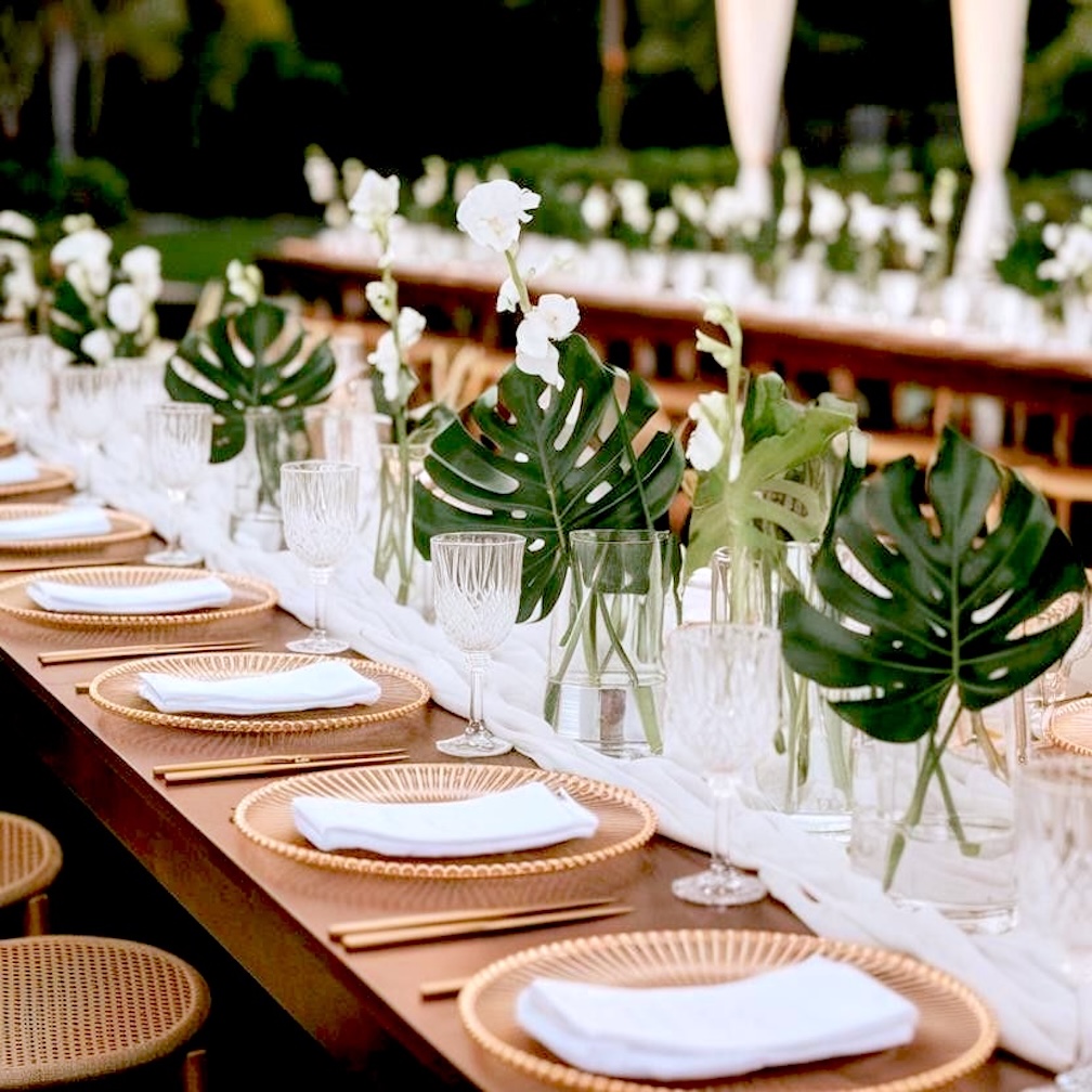 A row of place settings set up on a long table, alongside vases with small palm leaves.